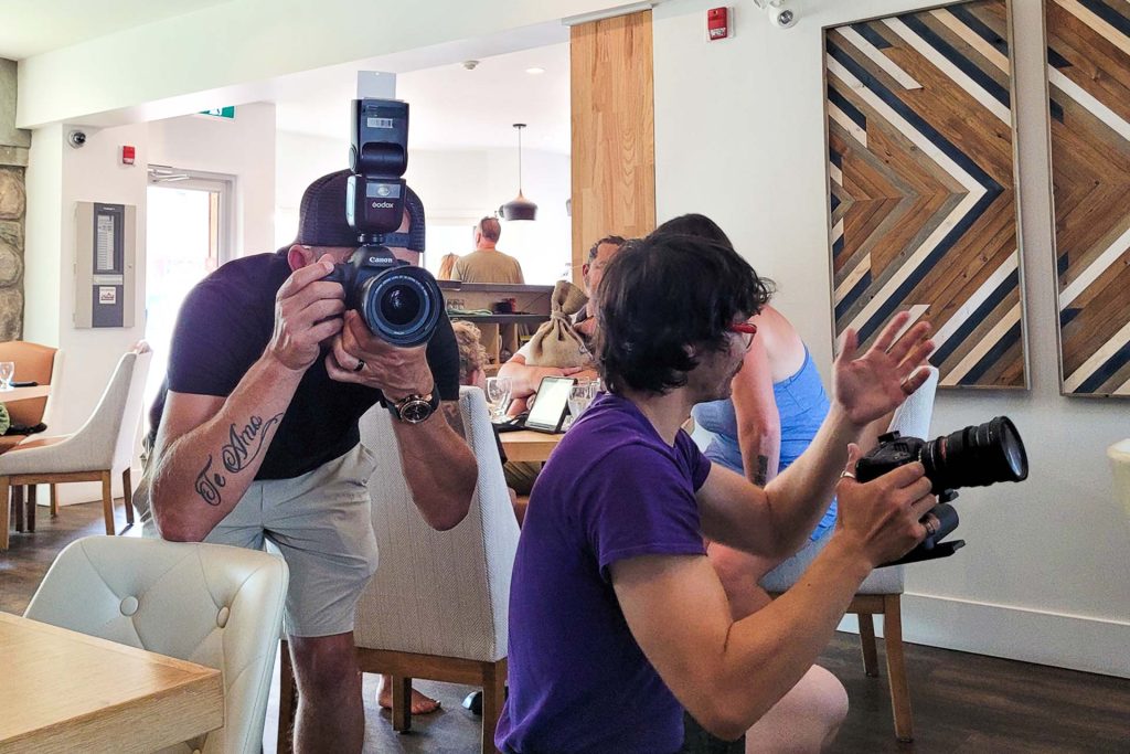 two men holding camera at a public event, there is a family sitting at the table beside them